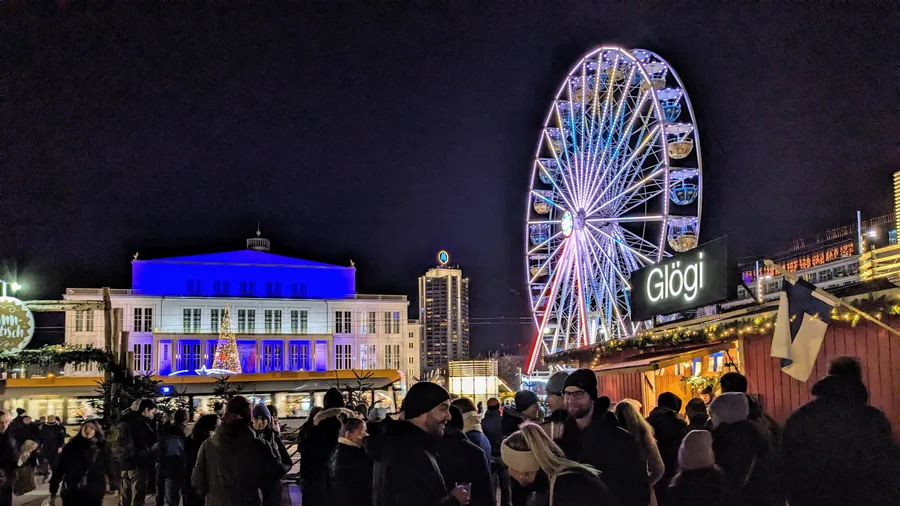 Weihnachtsmarkt mit beleuchtetem Riesenrad und Menschen vor einer Glühwein-Hütte