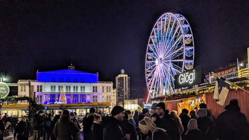 Weihnachtsmarkt mit beleuchtetem Riesenrad und Menschen vor einer Glühwein-Hütte